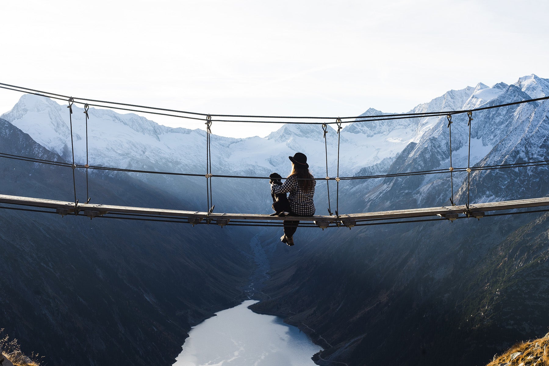 Person sitting on a suspension bridge with their dog, overlooking a mountain valley