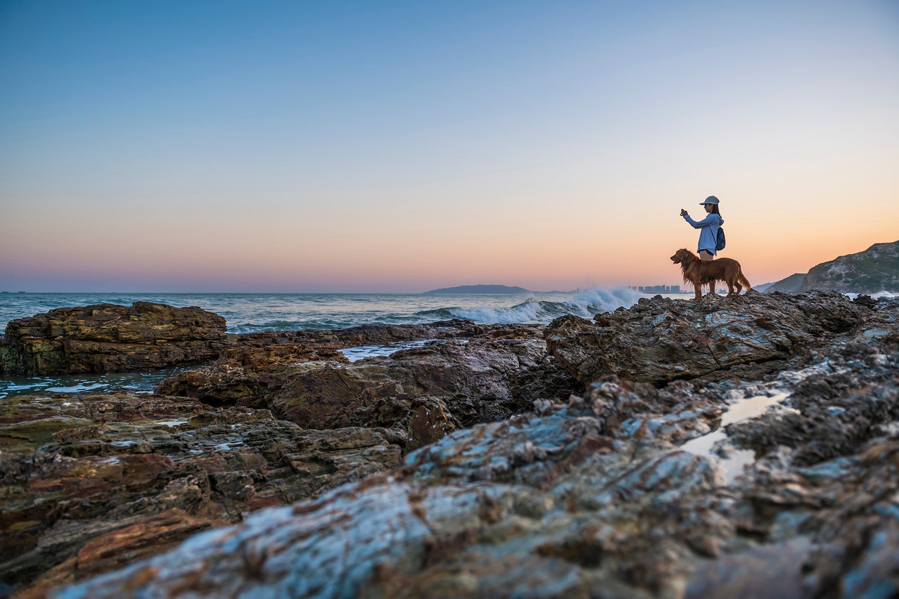 Person and dog standing on rocky beach at sunset
